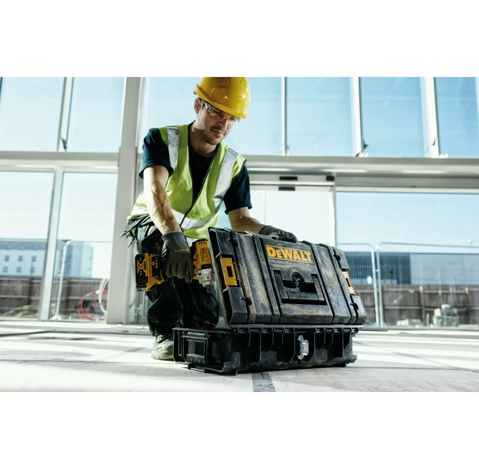 A person wearing a safety vest and yellow hard hat is handling a DEWALT toolbox on a construction site with large windows in the background.