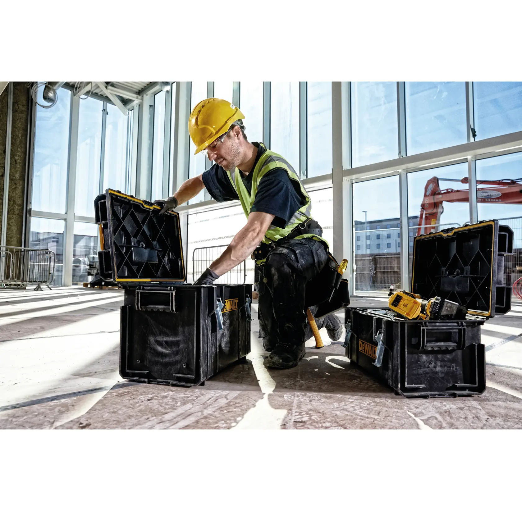 A construction worker wearing a yellow hard hat and safety vest is kneeling indoors, opening a black DEWALT tool box with other DEWALT tool boxes and tools nearby on the floor. The area has large windows and building equipment is visible outside.