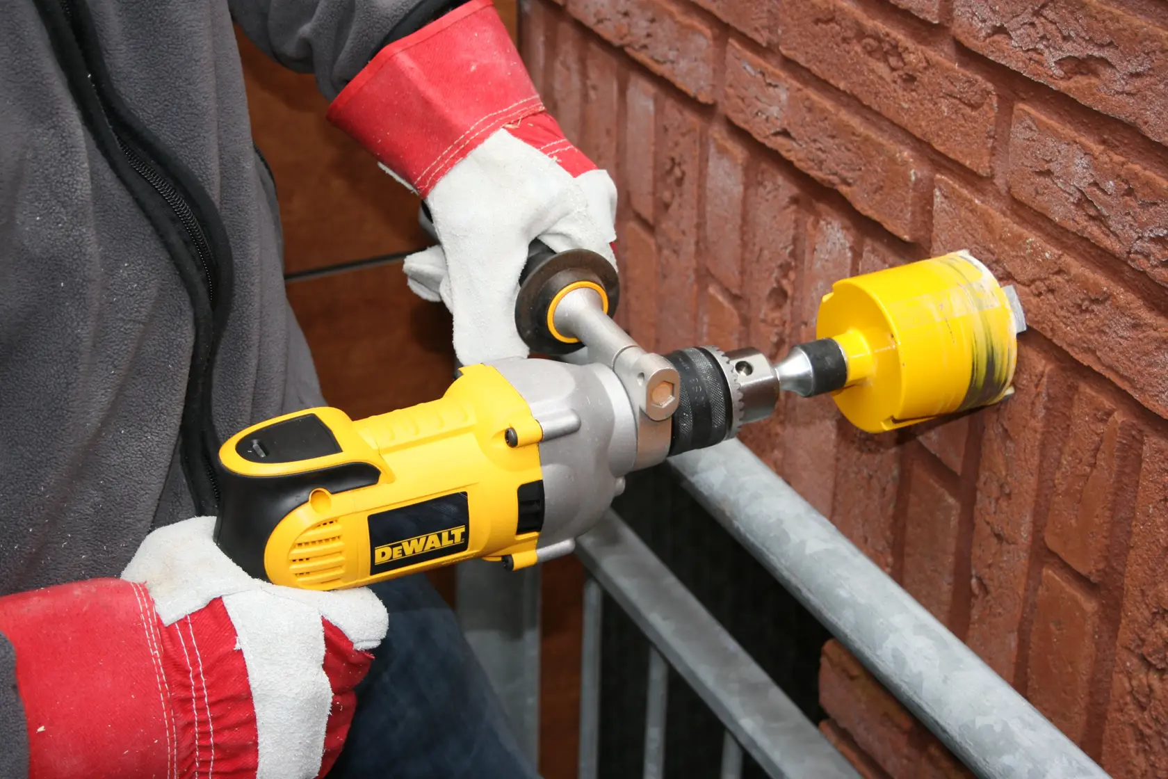 A person wearing protective gloves is using a yellow DEWALT power drill with a hole saw attachment to drill into a brick wall.
