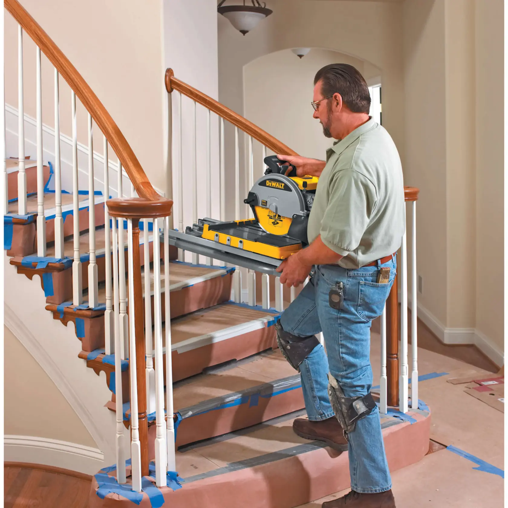 A person wearing knee pads carries a DEWALT D24000 tile saw up a staircase. The stairs are covered with protective paper and blue tape. The setting appears to be indoors during a renovation.