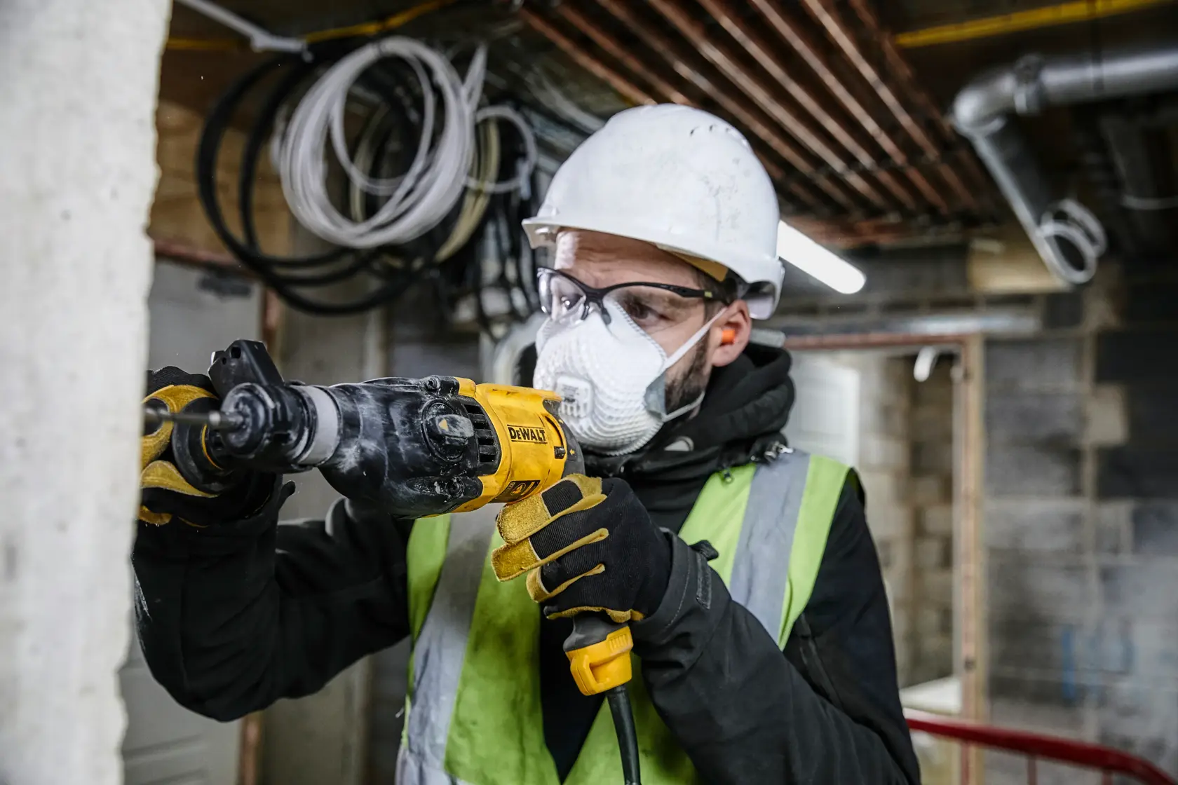 A person wearing a safety helmet, gloves, and a high-visibility vest is using a DEWALT power drill to work on a concrete wall in a construction environment. The face is blurred for privacy.