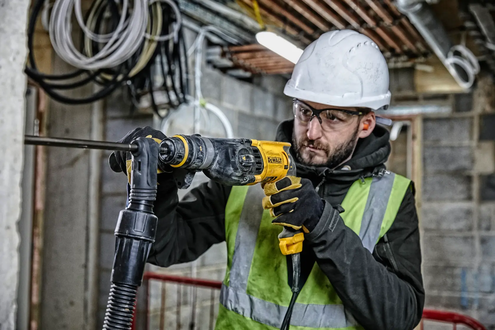 A person wearing a safety helmet, gloves, and a high-visibility vest is using a DEWALT D25134K rotary hammer drill on a construction site. The background shows exposed cables and piping in an unfinished building.