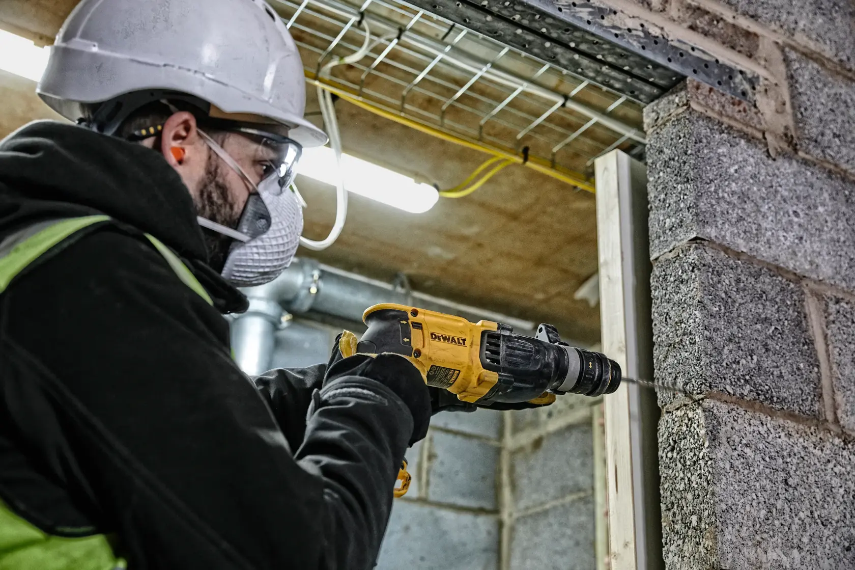 A worker wearing a safety helmet and gloves is using a DEWALT D25134K rotary hammer to drill into a concrete wall at a construction site.