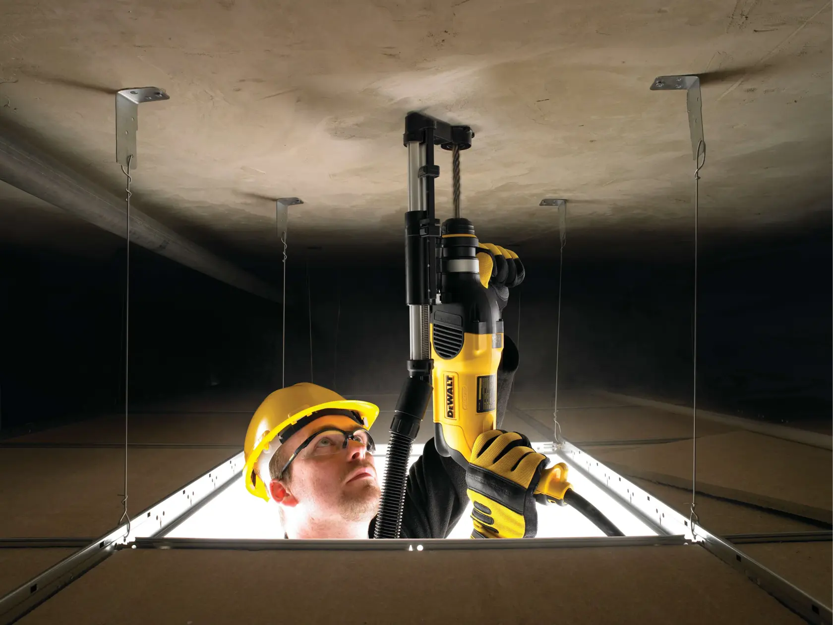 A person wearing safety gear and gloves uses a yellow DEWALT power tool to drill into a ceiling inside a construction site, viewed from an opening in the ceiling tiles.