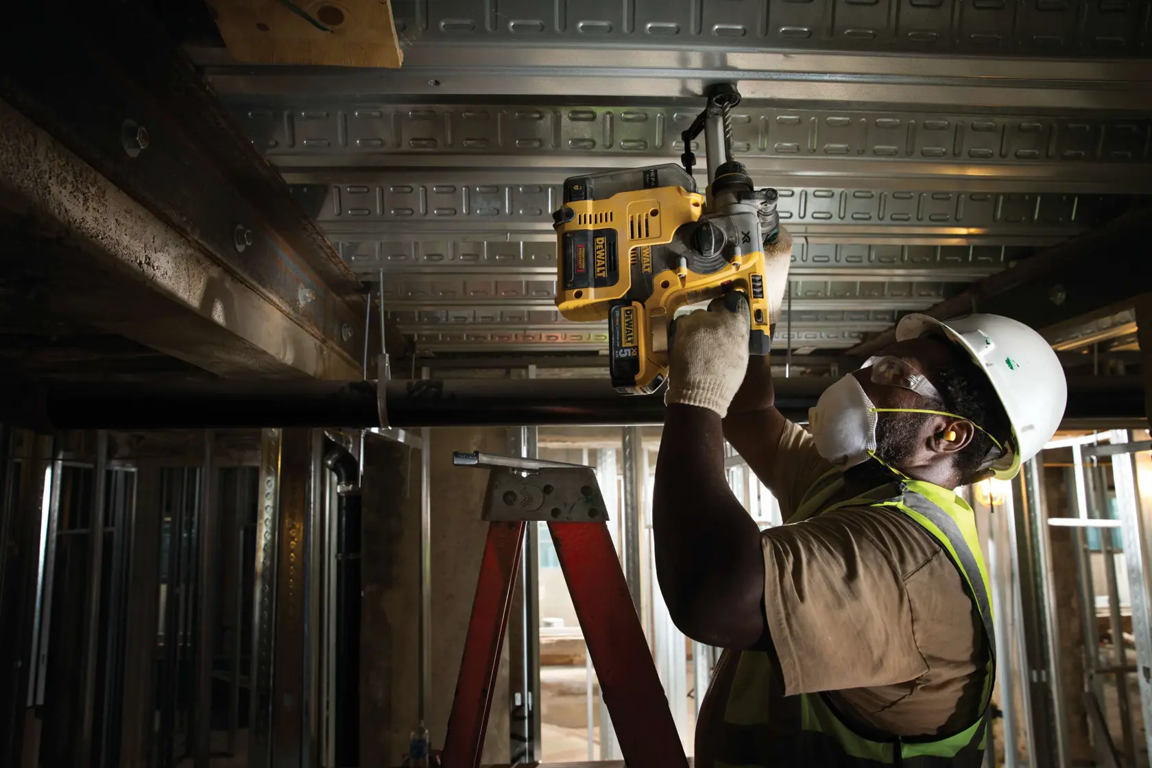 Ein Bauarbeiter mit Sicherheitsweste, Handschuhen, Helm und Atemmaske verwendet einen gelben DEWALT-Bohrhammer, um über Kopf an einer Metalldeckenstruktur in einem Gebäude zu arbeiten.