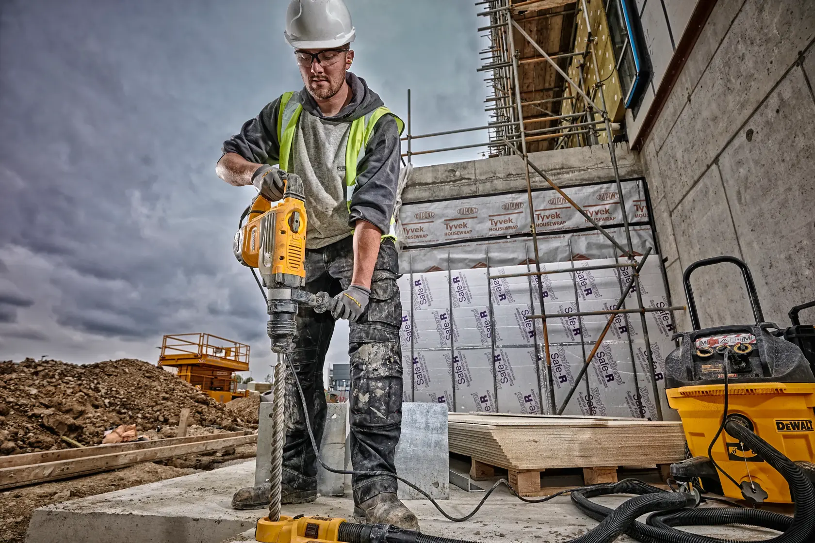 A construction worker wearing a safety helmet and high-visibility vest operates a DEWALT rotary hammer drill (SKU: D25733K) on a building site. The worker's face is blurred, and there is construction equipment, scaffolding, and insulation materials visible in the background.