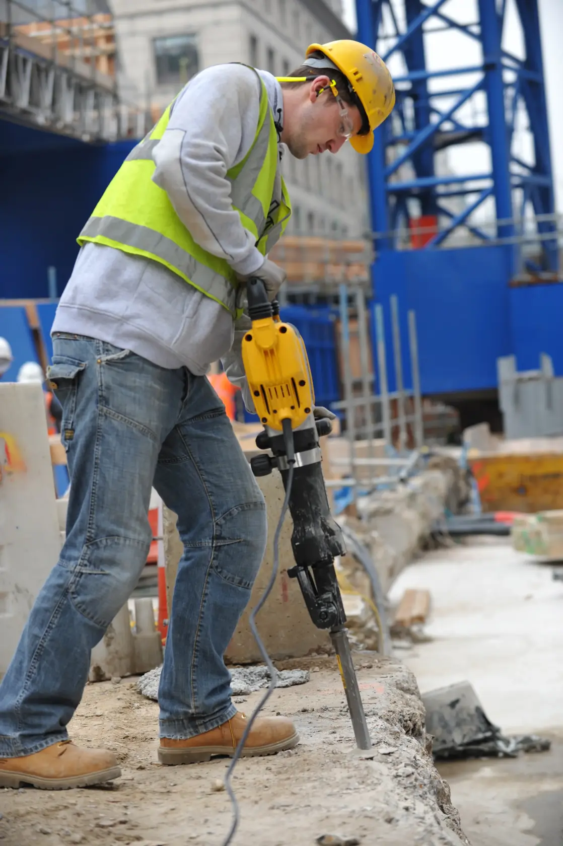 Worker uses a DEWALT D25960K demolition hammer on concrete at a construction site, wearing safety gear.