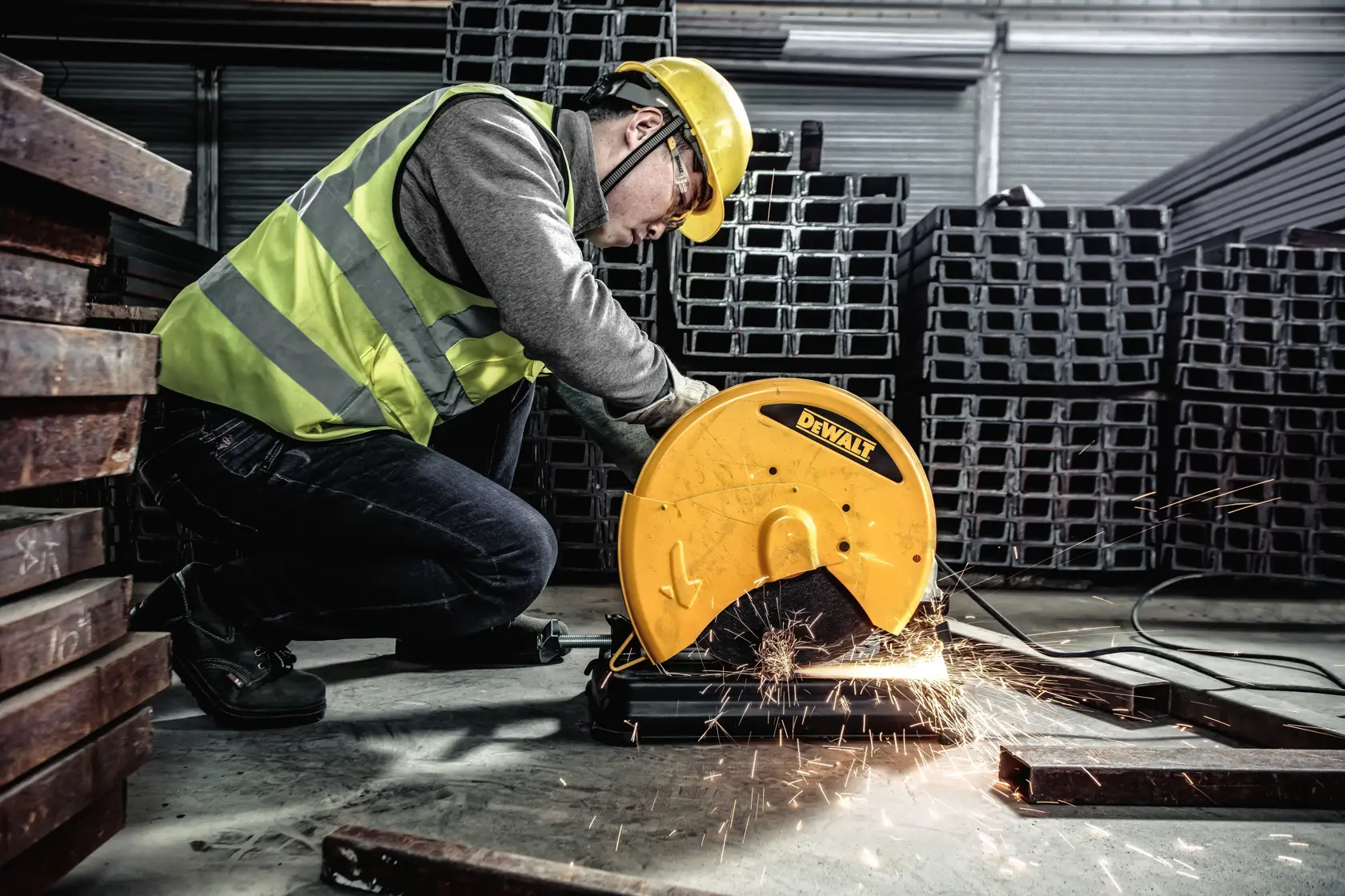 A person wearing a yellow safety helmet and high-visibility vest operates a DEWALT chop saw, cutting metal pieces in an industrial warehouse with metal beams stacked in the background.