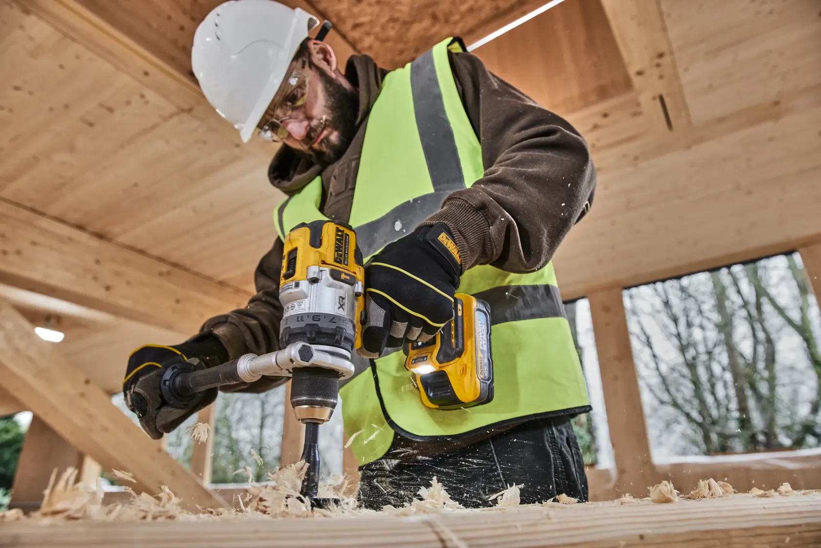 Person using a DEWALT cordless drill to bore wood, wearing safety gear and high-visibility vest on a construction site.