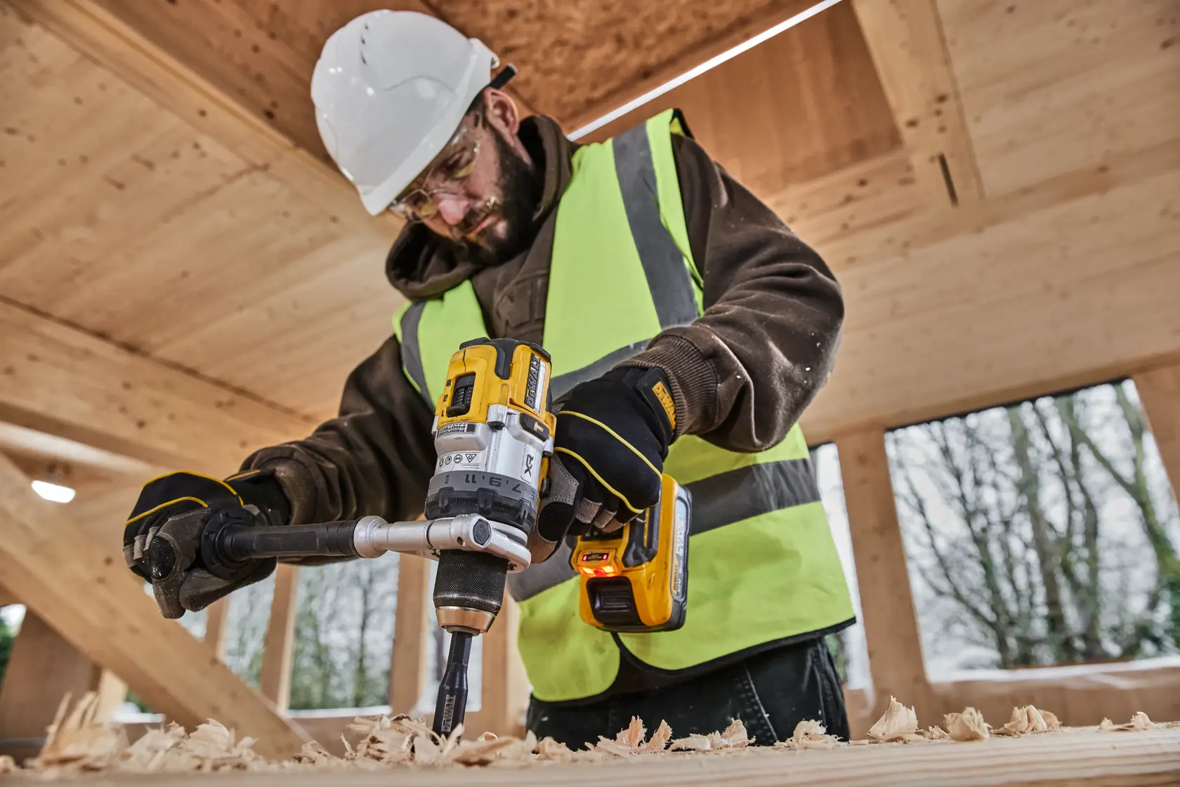Person uses a DEWALT cordless drill on wood, wearing safety gear and a high-visibility vest at a construction site.