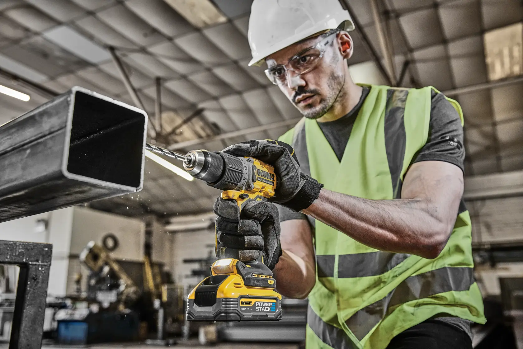 Worker using a DEWALT drill on a metal beam in an industrial setting, wearing safety gear and gloves.