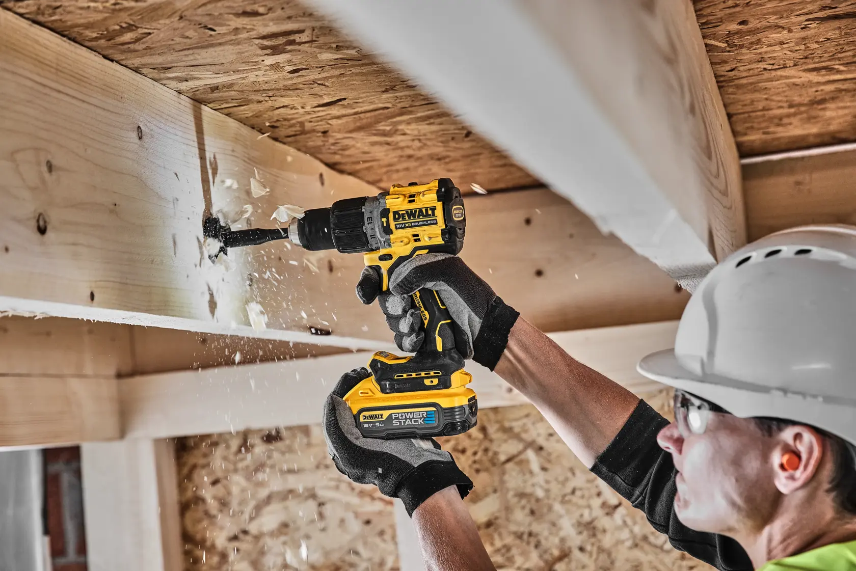 Person uses DEWALT cordless drill to make a hole in a wooden beam at a construction site, wood chips flying.