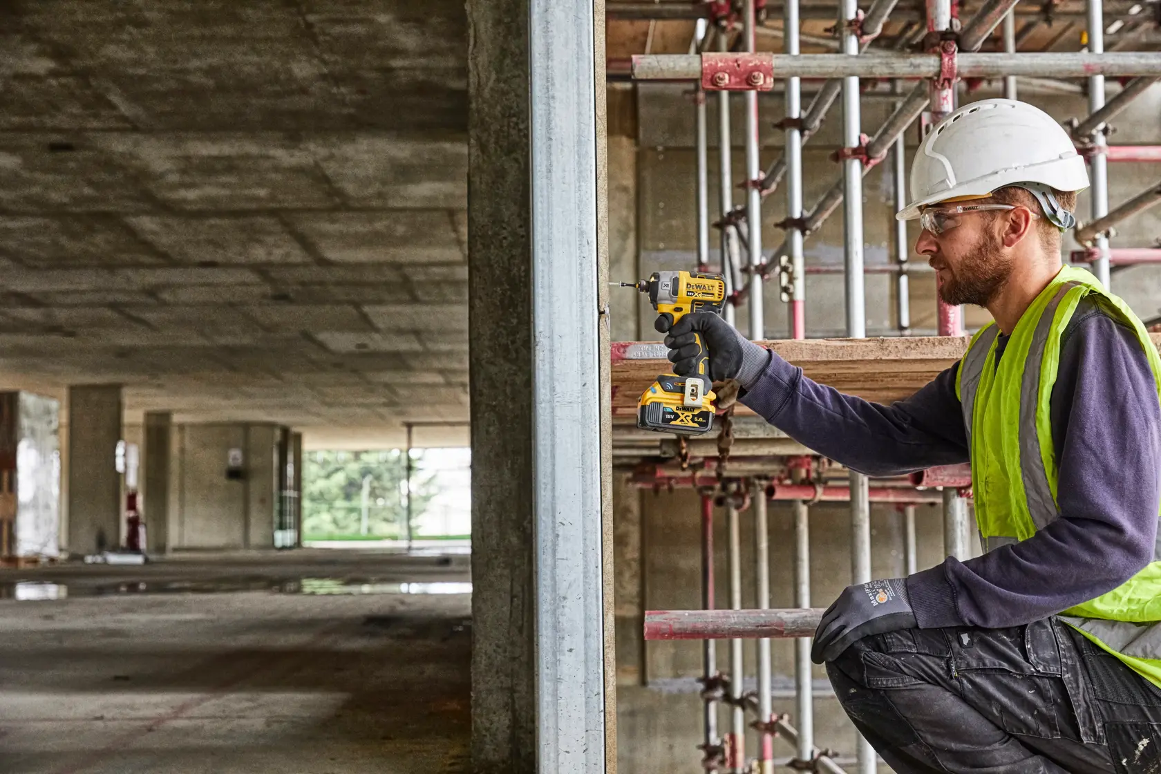 Worker uses a DEWALT drill to fasten metal on a construction site, wearing safety gear and high visibility vest.