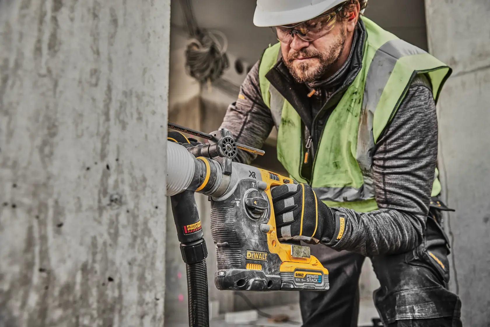 A construction worker using a DEWALT DCH273H2T rotary hammer drill with dust extraction, drilling into concrete. Worker is wearing safety gloves, high-visibility vest, and hard hat. The drill is branded DEWALT and features the PowerStack battery.