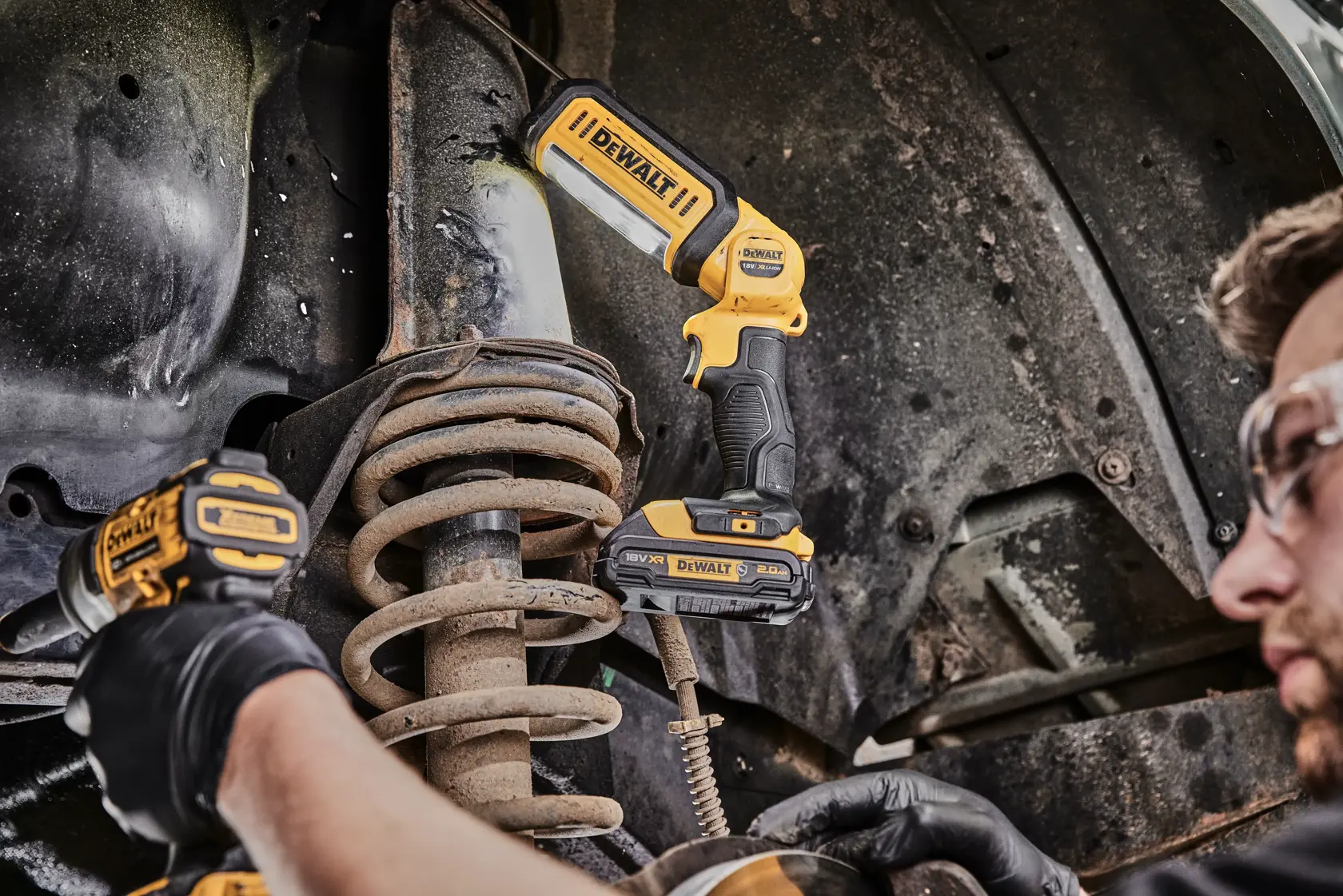 Eine Person kniet und arbeitet mit Werkzeugen an einer offenen elektrischen Einheit. Auf dem Boden steht eine tragbare DEWALT DCL050 LED-Arbeitsleuchte, die den Arbeitsbereich beleuchtet.