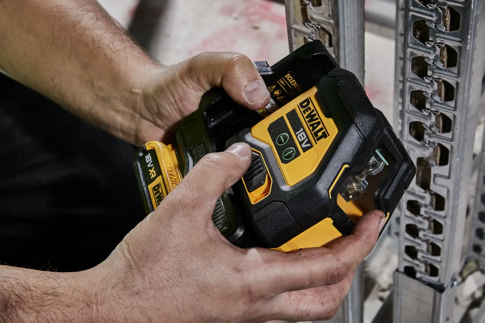 Hands adjusting a DEWALT 18V XR laser level near a metal framework at a construction site.