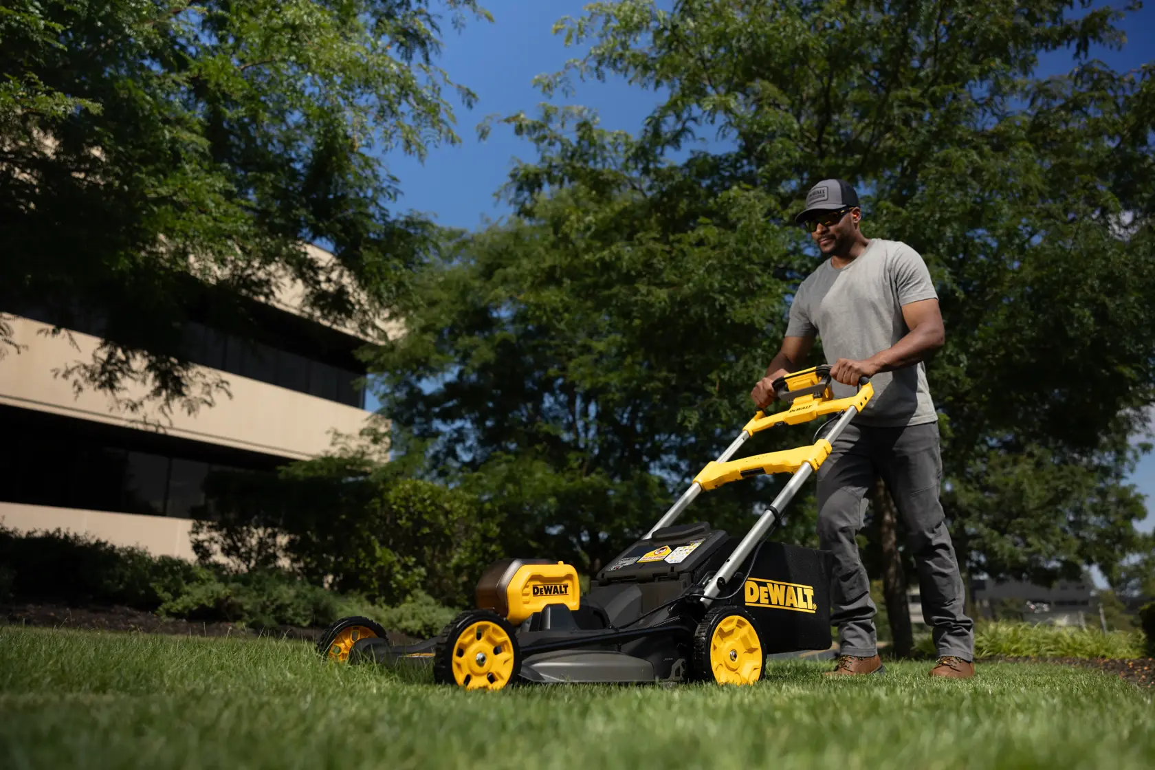 Persona usa una cortadora de césped DEWALT en exteriores, con edificios y árboles de fondo.
