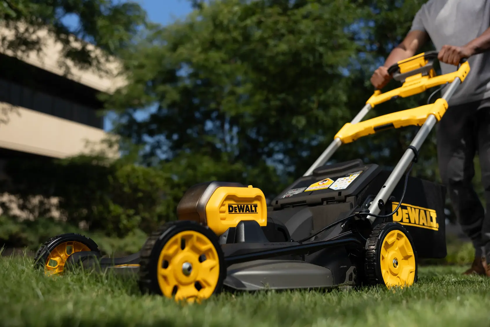 Una cortadora DEWALT se empuja sobre césped al aire libre, con árboles y edificio de fondo.