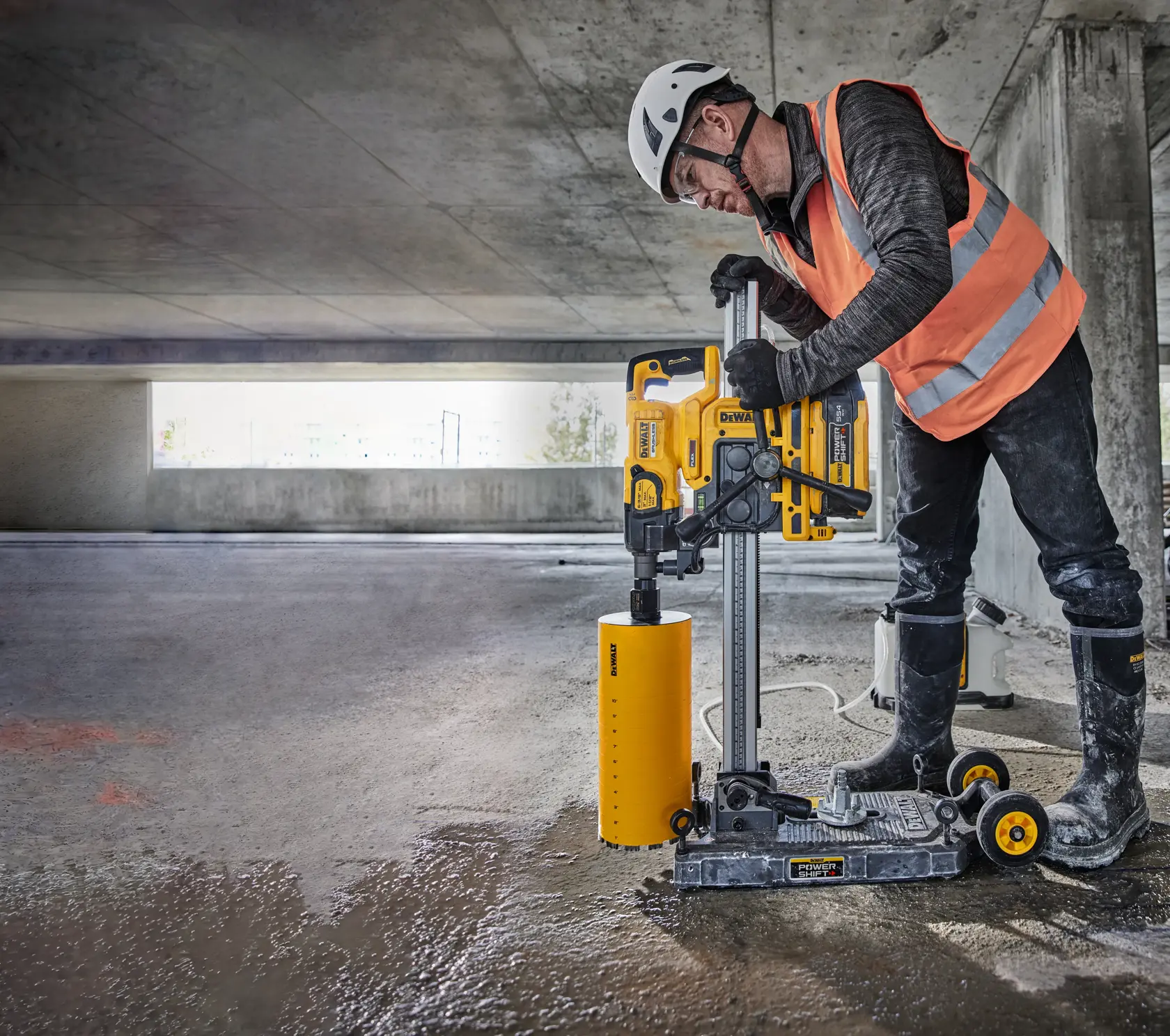 Ein Bauarbeiter mit Sicherheitsweste, Handschuhen und Helm hält ein DEWALT-Werkzeug mit Rädern auf einer Baustelle. Der Arbeiter steht vor einem LKW mit DEWALT-Branding und weiterem Equipment im Hintergrund.