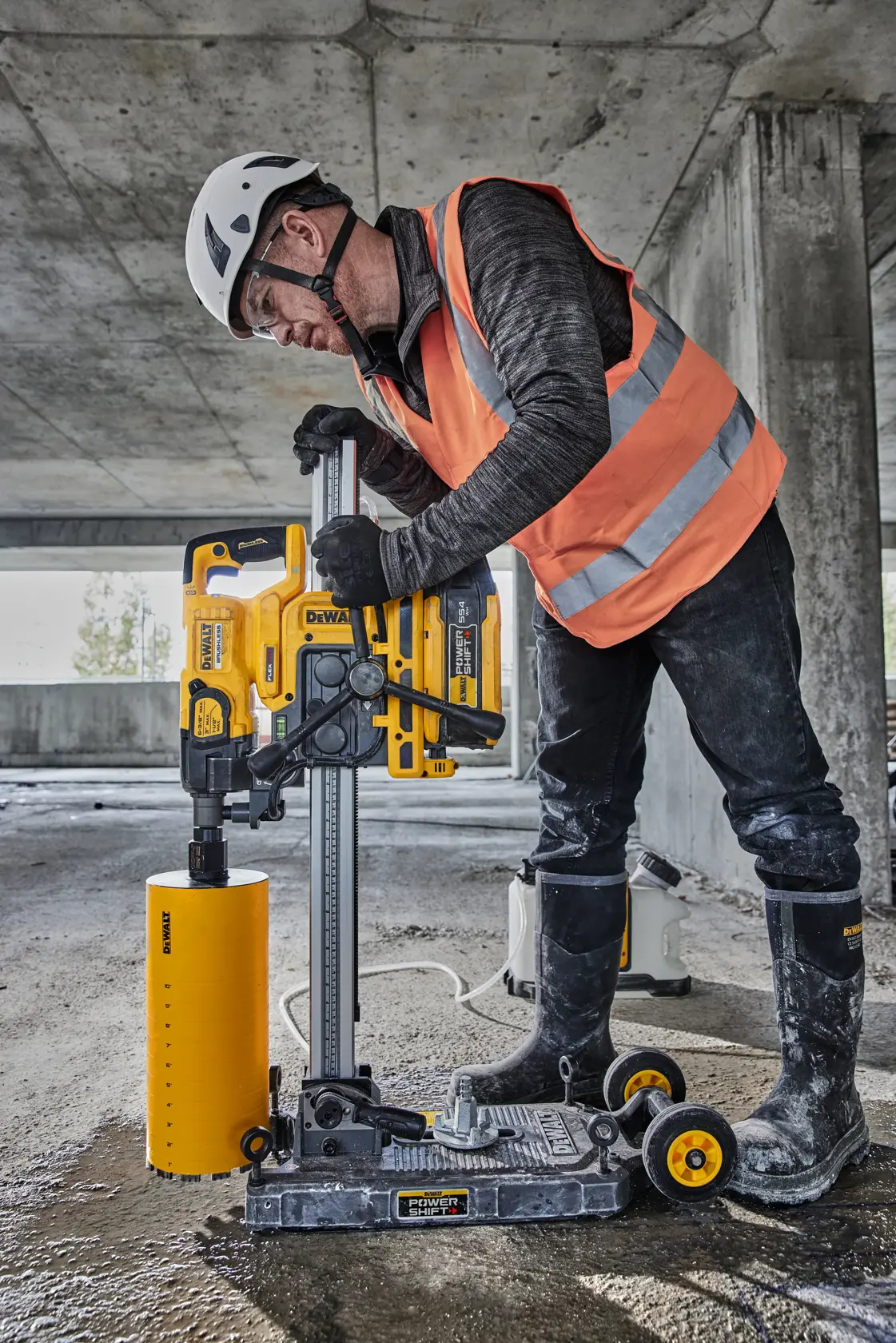 Eine Person mit Schutzhelm, Handschuhen und reflektierender Weste verwendet ein DEWALT Elektrowerkzeug auf einer Baustelle. Im Hintergrund sind DEWALT Werkzeugkasten und Geräte zu sehen.