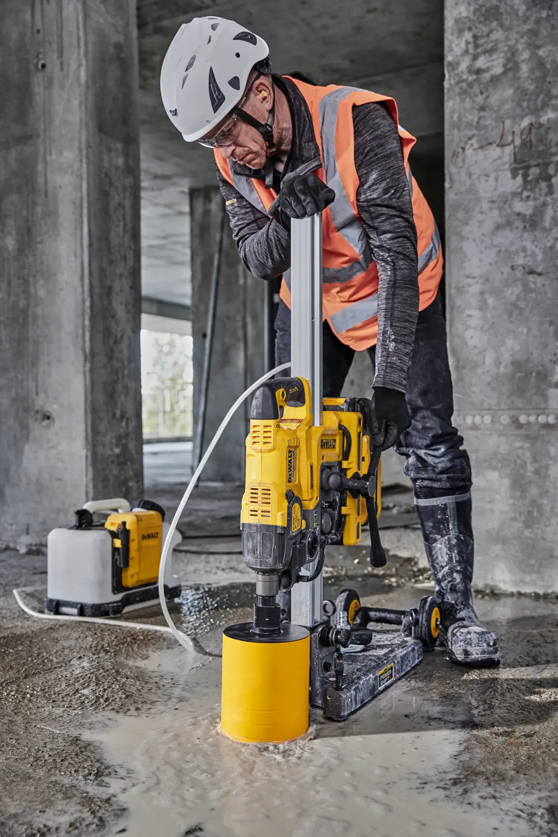 Ein Bauarbeiter mit Schutzhelm, Handschuhen und orangefarbener reflektierender Weste trägt ein DEWALT Werkzeug mit Rädern und einem gelben Teil auf der Schulter und geht an einer Betonwand vorbei.