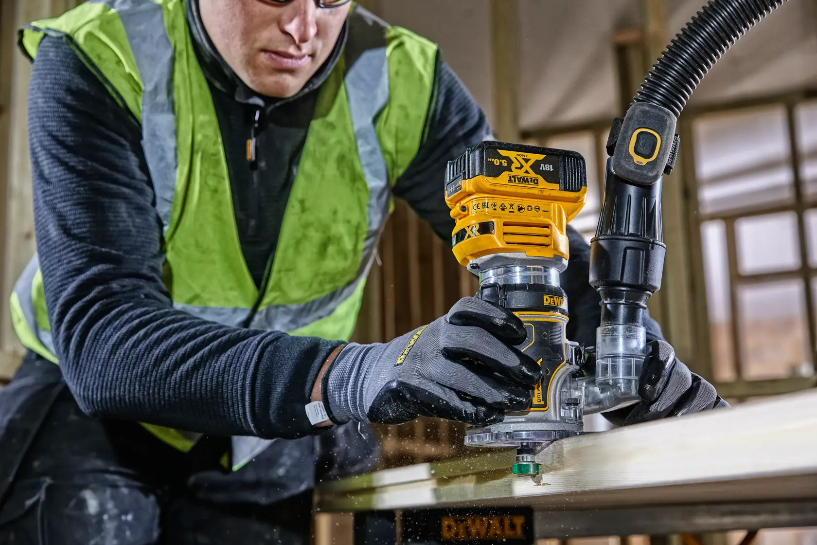 Worker uses a DEWALT DCW604 router on wood, wearing gloves and a safety vest in a workshop.