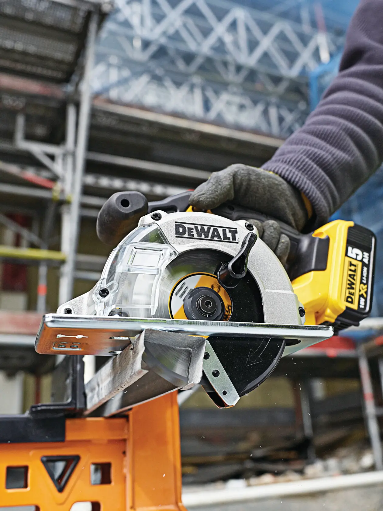 DEWALT cordless circular saw cutting metal outdoors, held by a gloved hand, construction site in background.