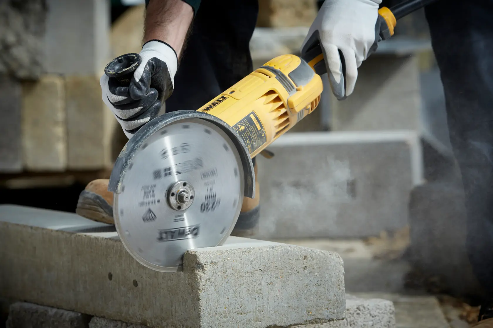 Person uses a DEWALT angle grinder with a diamond blade to cut a concrete block outdoors, wearing safety gloves.