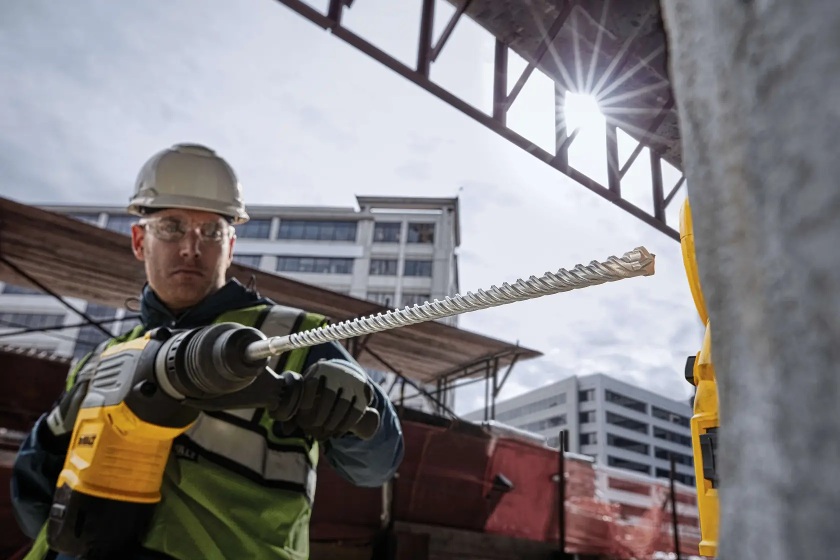 Persona usando taladro rotativo DEWALT amarillo en concreto, con botas y guantes, en obra.