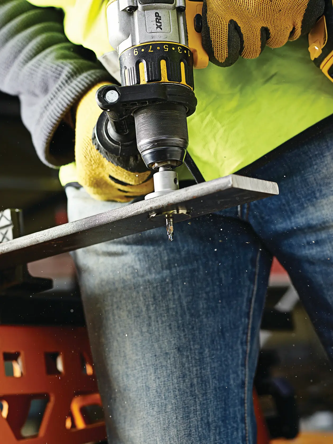 Person uses DEWALT drill to bore hole in metal sheet, wearing gloves and safety gear.