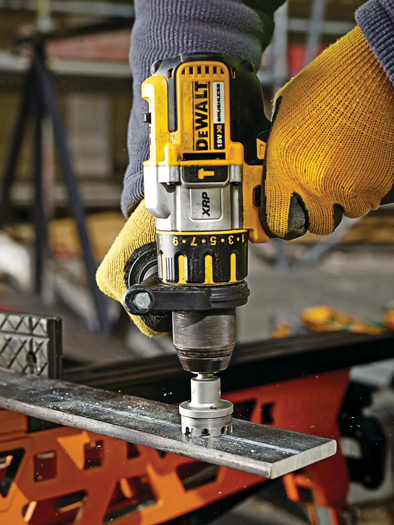 DEWALT cordless drill in gloved hands cutting a hole in a metal bar at a workshop.