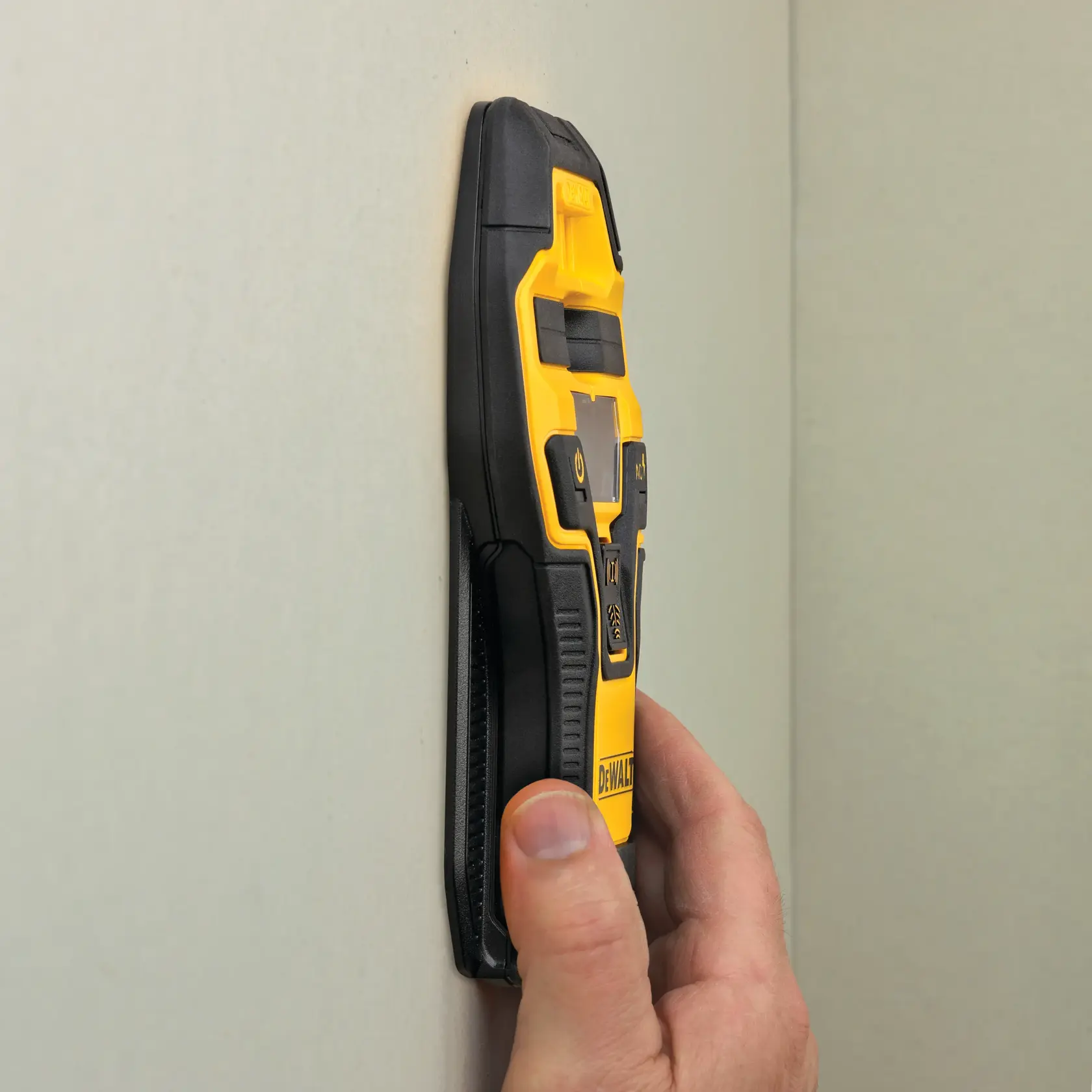 A hand holds a yellow and black DEWALT stud finder against a wall, demonstrating its use for detecting studs behind surfaces.
