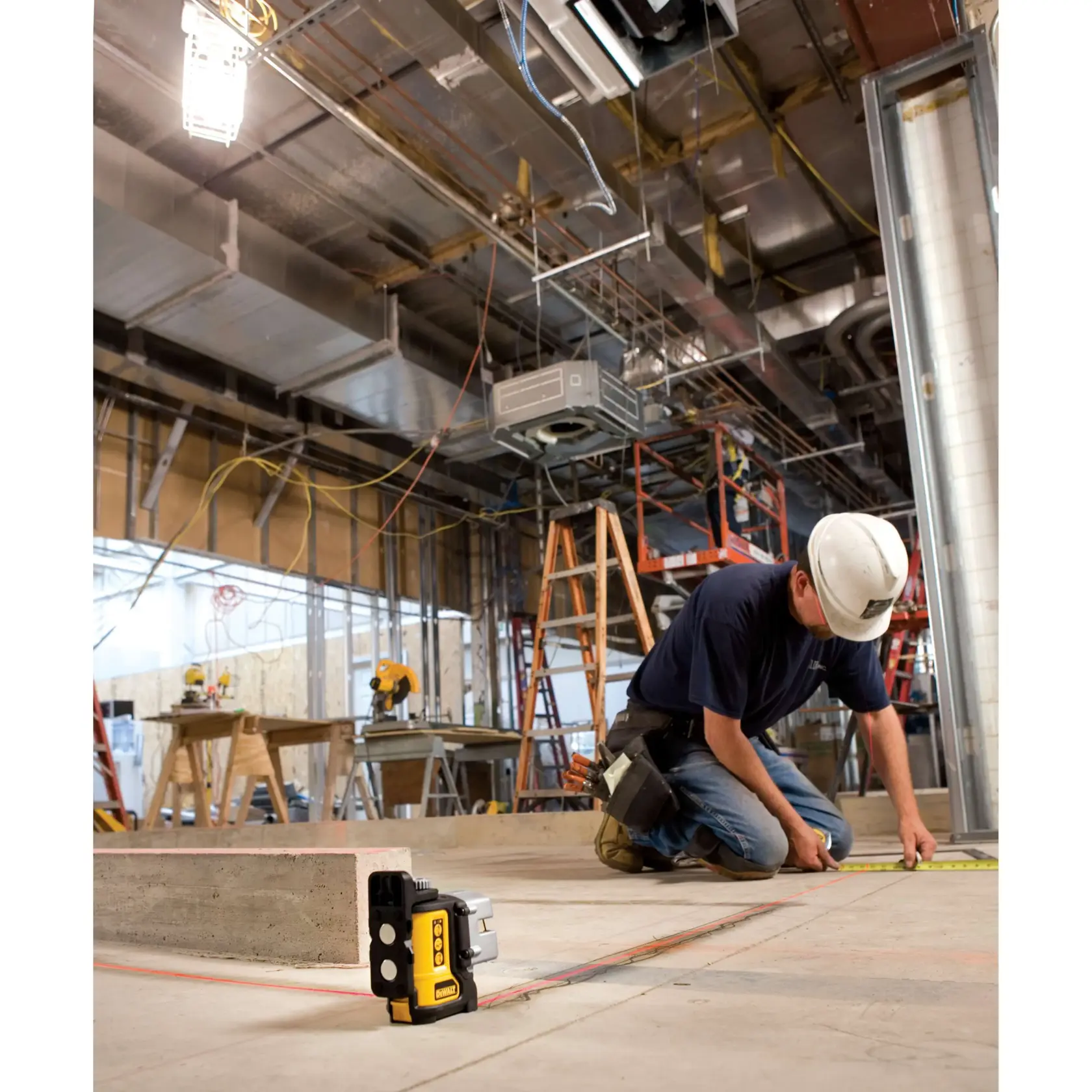Un niveau laser DEWALT DW089K est placé au sol sur un chantier de construction, tandis qu’un ouvrier portant un casque et une ceinture à outils mesure avec un mètre ruban sur le sol. L’arrière-plan montre divers équipements de construction, des échelles et des tuyaux du plafond exposés.
