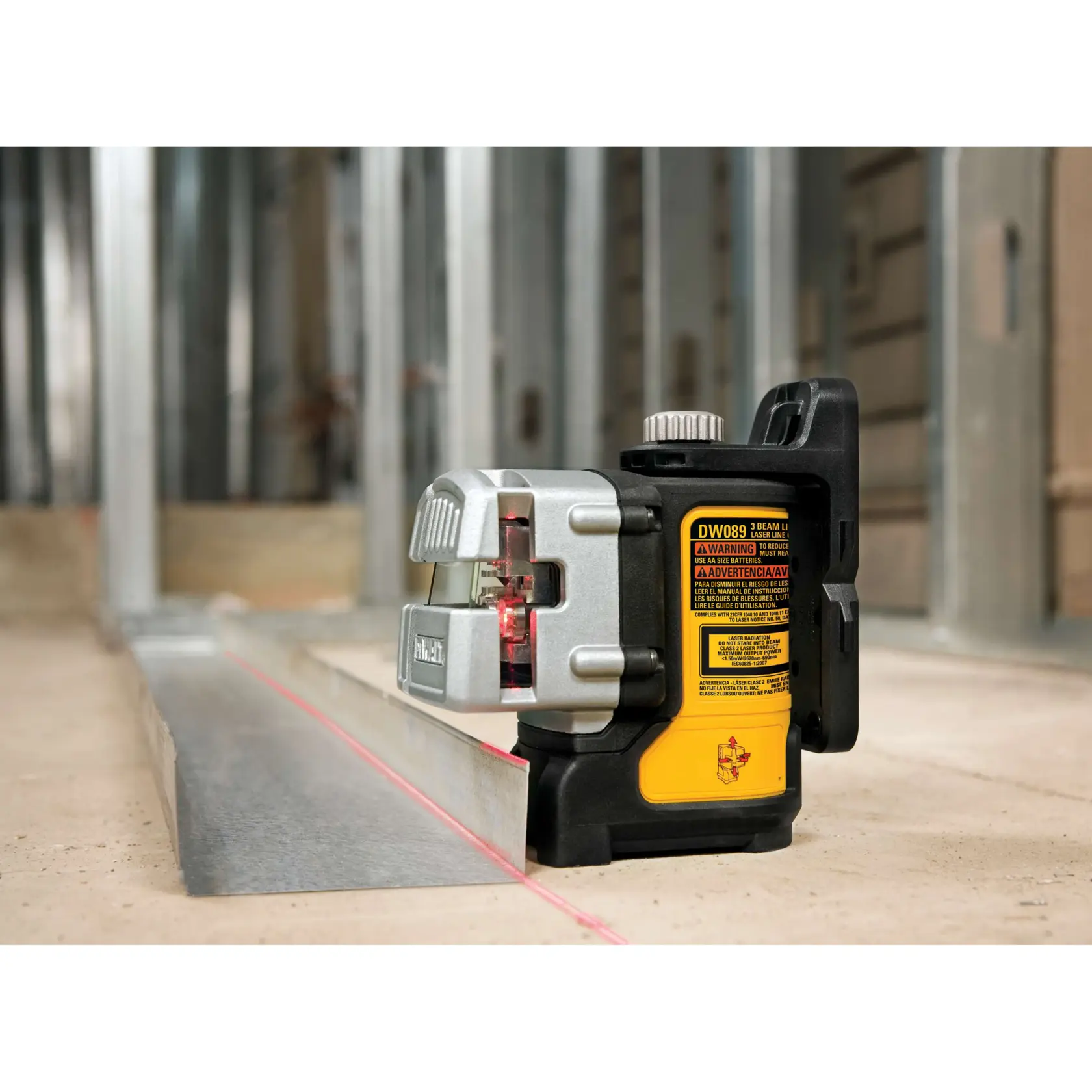 Niveau laser à trois faisceaux DEWALT DW089K posé sur le sol d'un chantier, projetant une ligne laser rouge le long d'un rail de structure métallique avec des montants muraux en métal en arrière-plan.