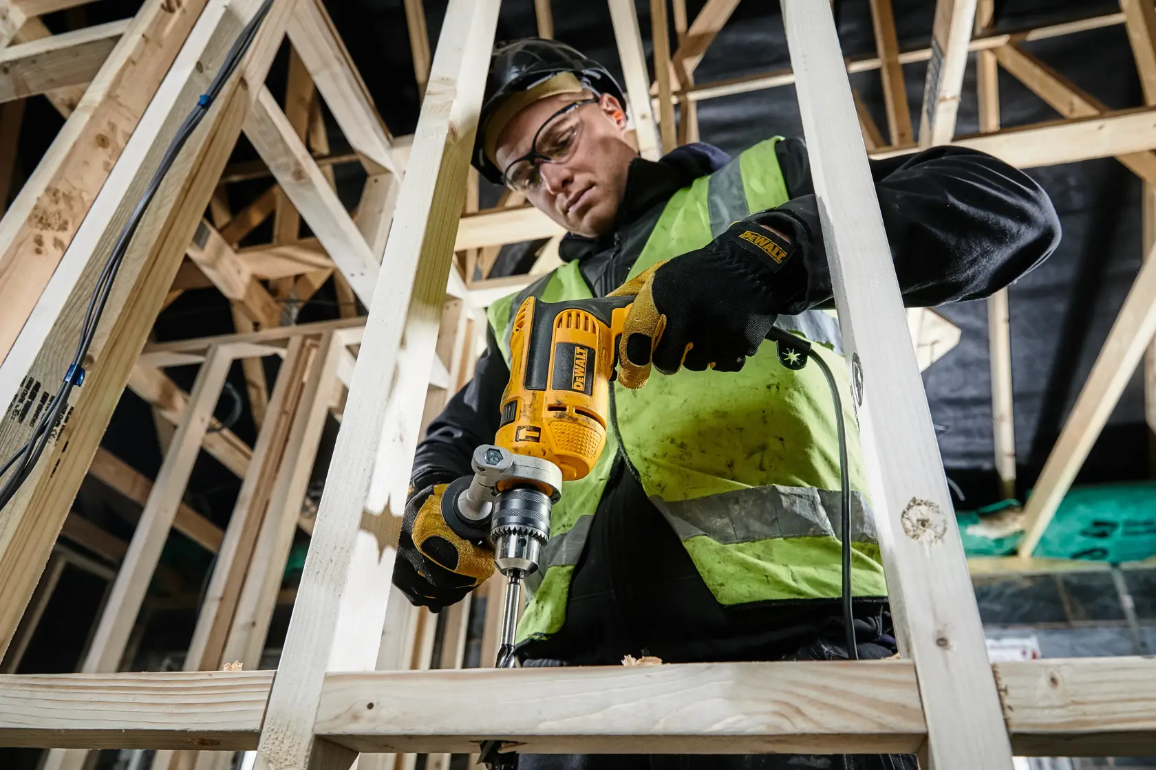 DEWALT DWD024K drill in use on wooden beams at a construction site by a person wearing safety gear.