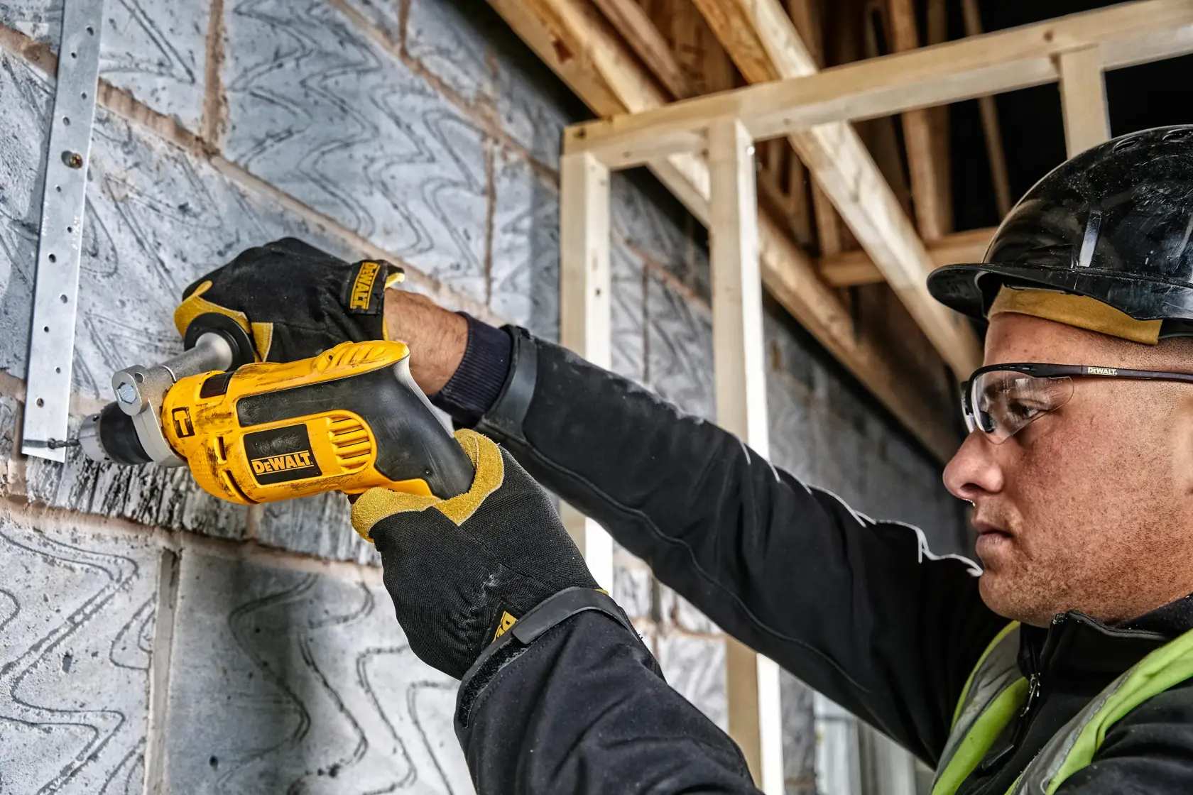 Gloved worker uses DEWALT drill DWD024K on a brick wall at a construction site. Face is blurred for privacy.
