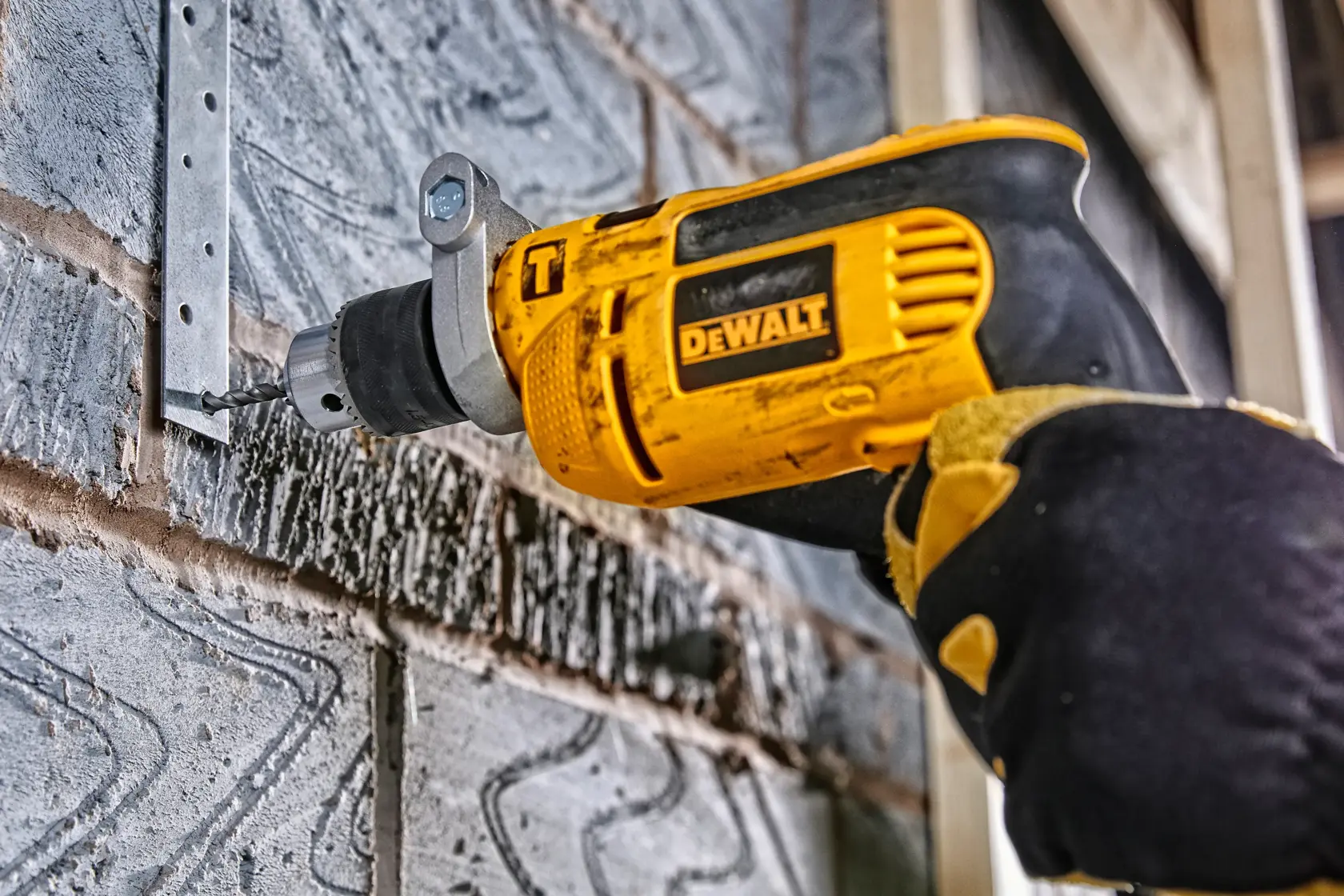 DEWALT drill in use, drilling into a metal bracket on a brick wall. Gloved hand holds the yellow power tool.