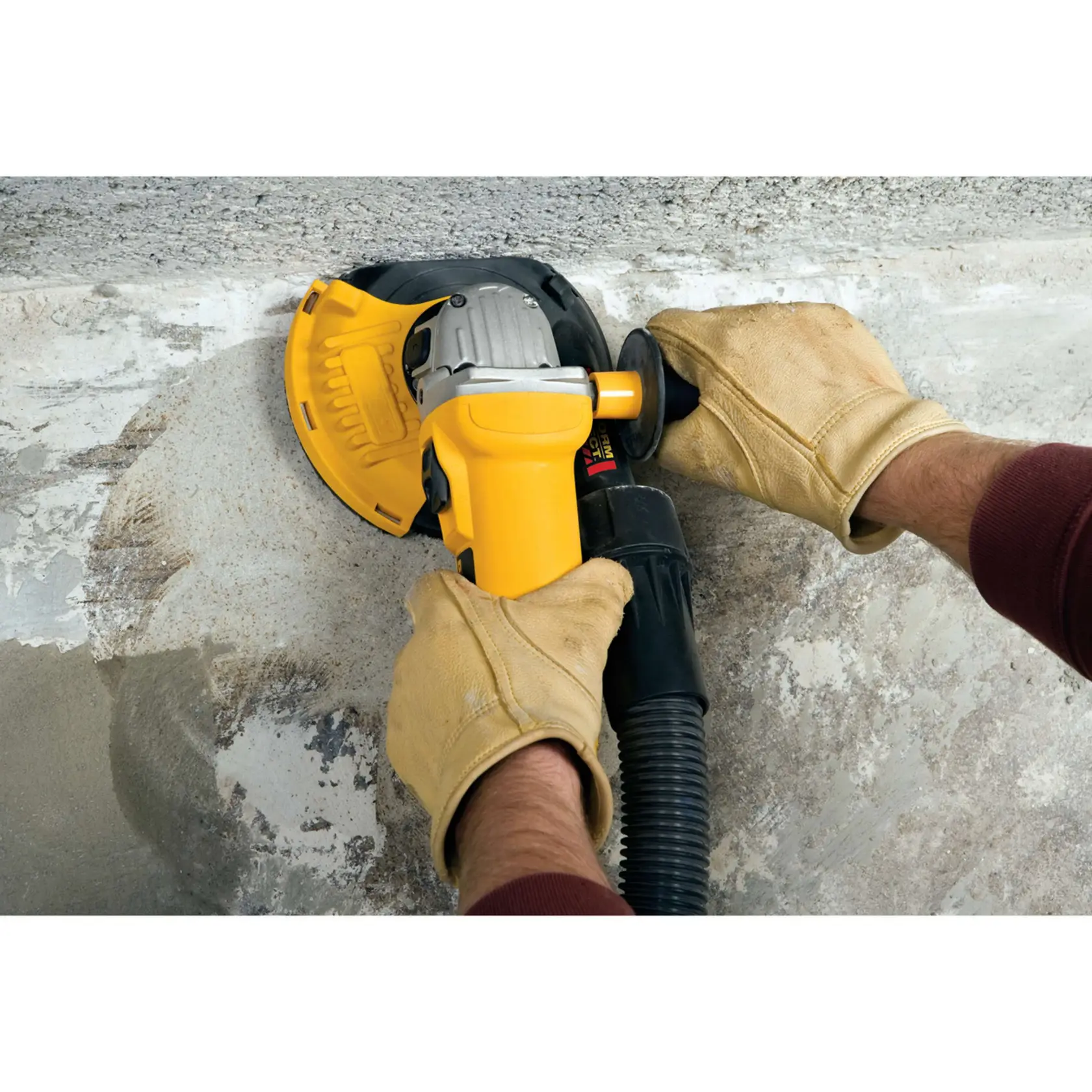 Ein Bauarbeiter mit gelbem Schutzhelm, Handschuhen und Gesichtsmaske benutzt ein DEWALT DWE46150 Oberflächen-Schleifwerkzeug auf einem Betonboden, während im Hintergrund ein gelber DEWALT Staubabsaugungsstaubsauger zu sehen ist. Die Szene spielt in einer industriellen oder baulichen Umgebung.