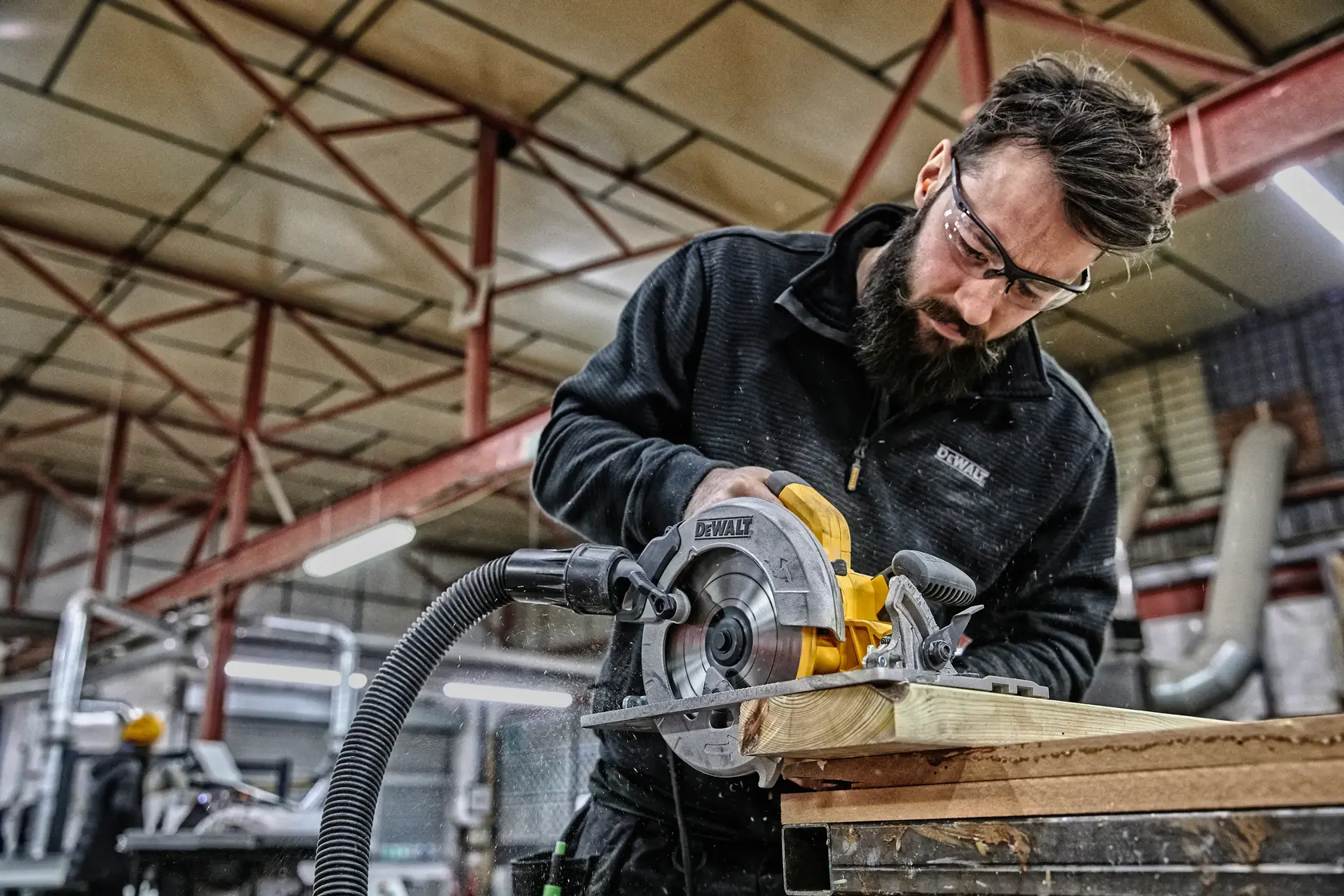 Worker using DEWALT DWE576K circular saw to cut wood in an industrial workshop, dust extraction hose attached.
