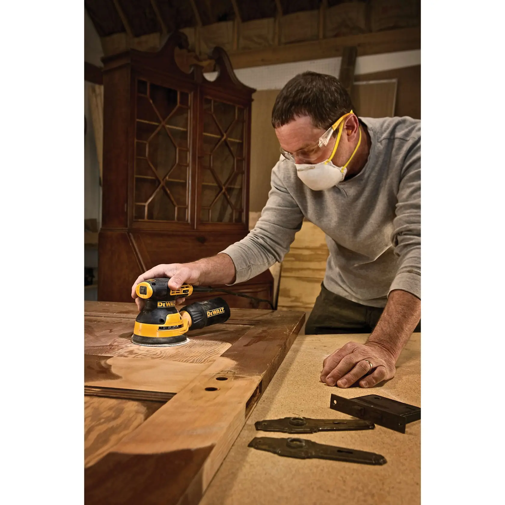 A person uses a DEWALT DWE6423 random orbital sander to smooth a large wooden surface in a workshop. The person’s face is blurred for privacy. Woodworking tools and cabinet parts are visible on the workspace.