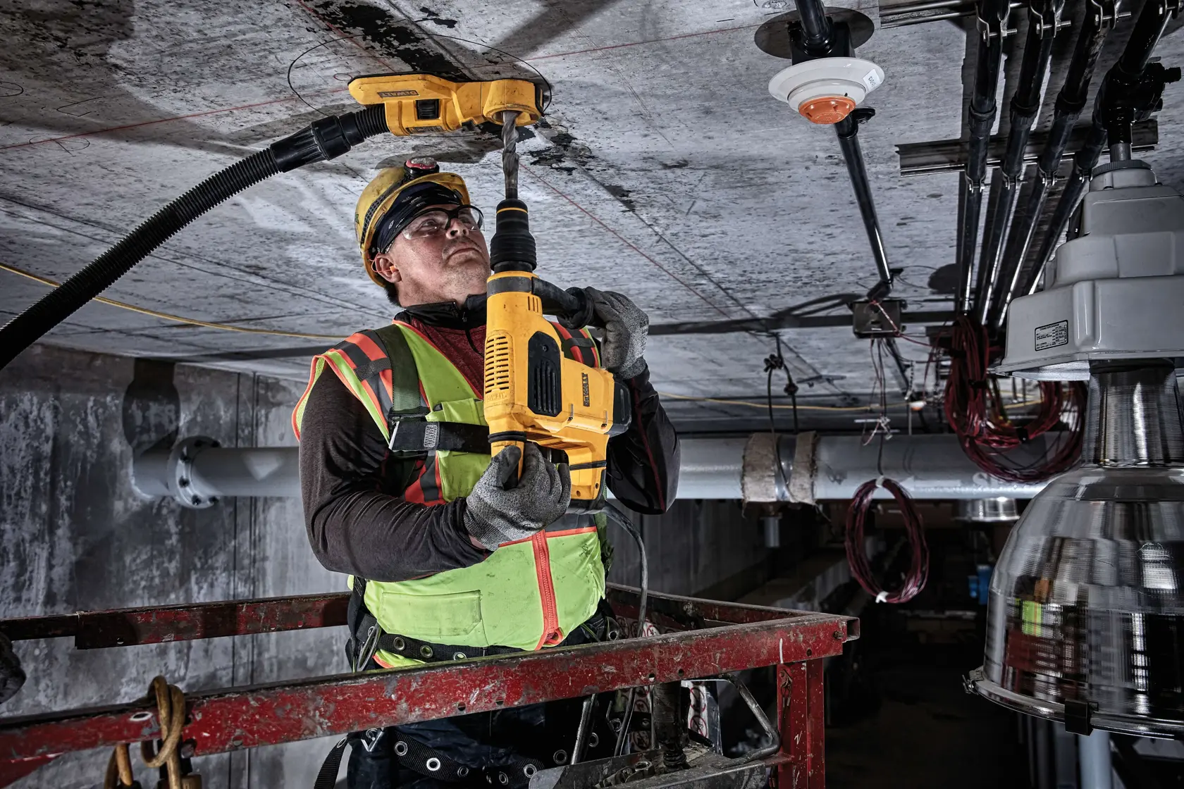 Ein Bauarbeiter mit Sicherheitsausrüstung verwendet ein DEWALT DWH050 Werkzeug, um eine Decke in einer industriellen Umgebung zu bohren. Der Arbeiter steht auf einer Hebebühne und eine Staubabsaugung ist an das Werkzeug angeschlossen.