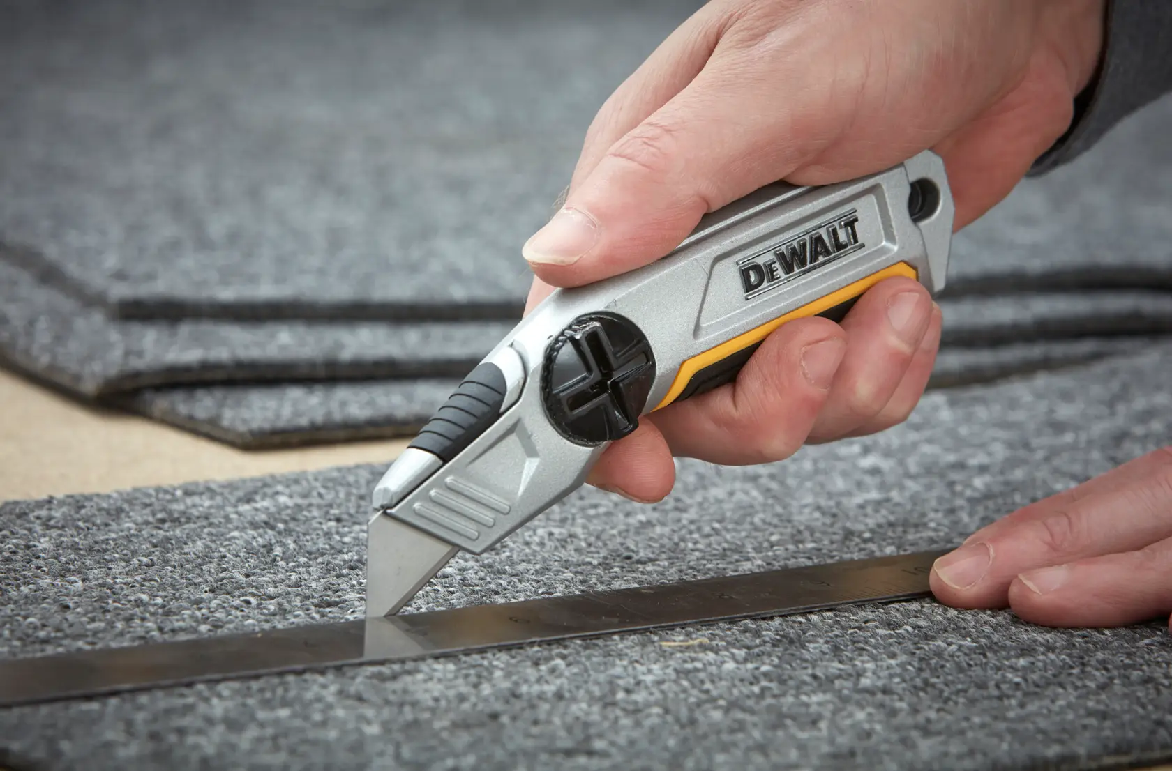 Hand using a DEWALT utility knife to cut gray carpet along a metal ruler.