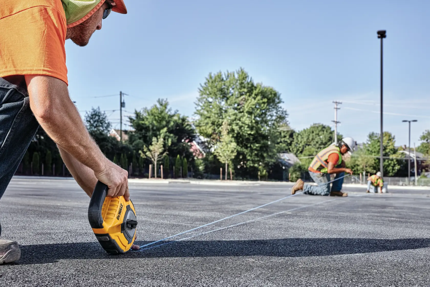 A person uses a DEWALT chalk reel to mark a straight line on asphalt, with two other workers in safety vests and hard hats working in the background on the same surface outdoors.