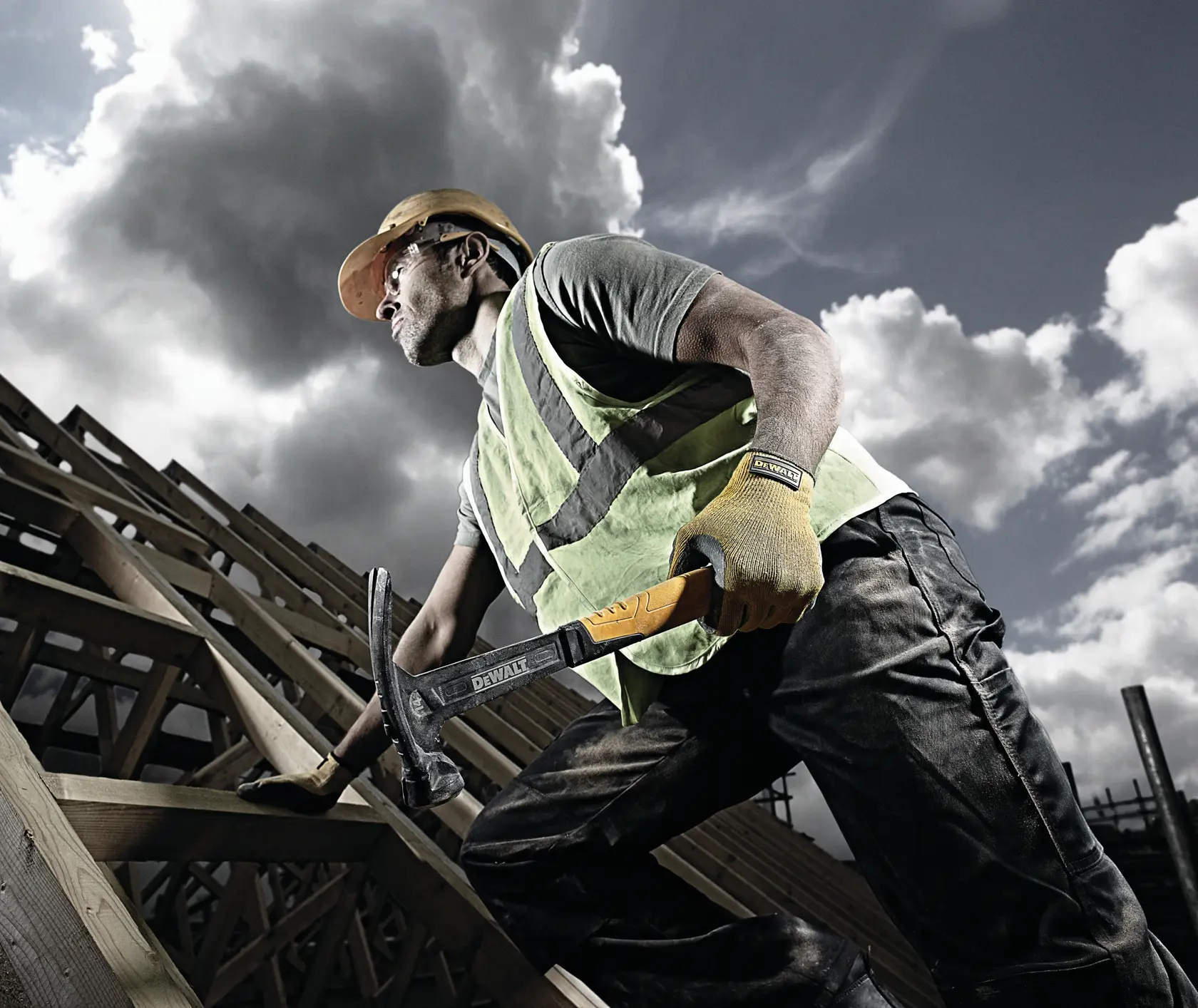 Worker in safety gear using a DEWALT hammer on a wooden roof structure, cloudy sky in the background.