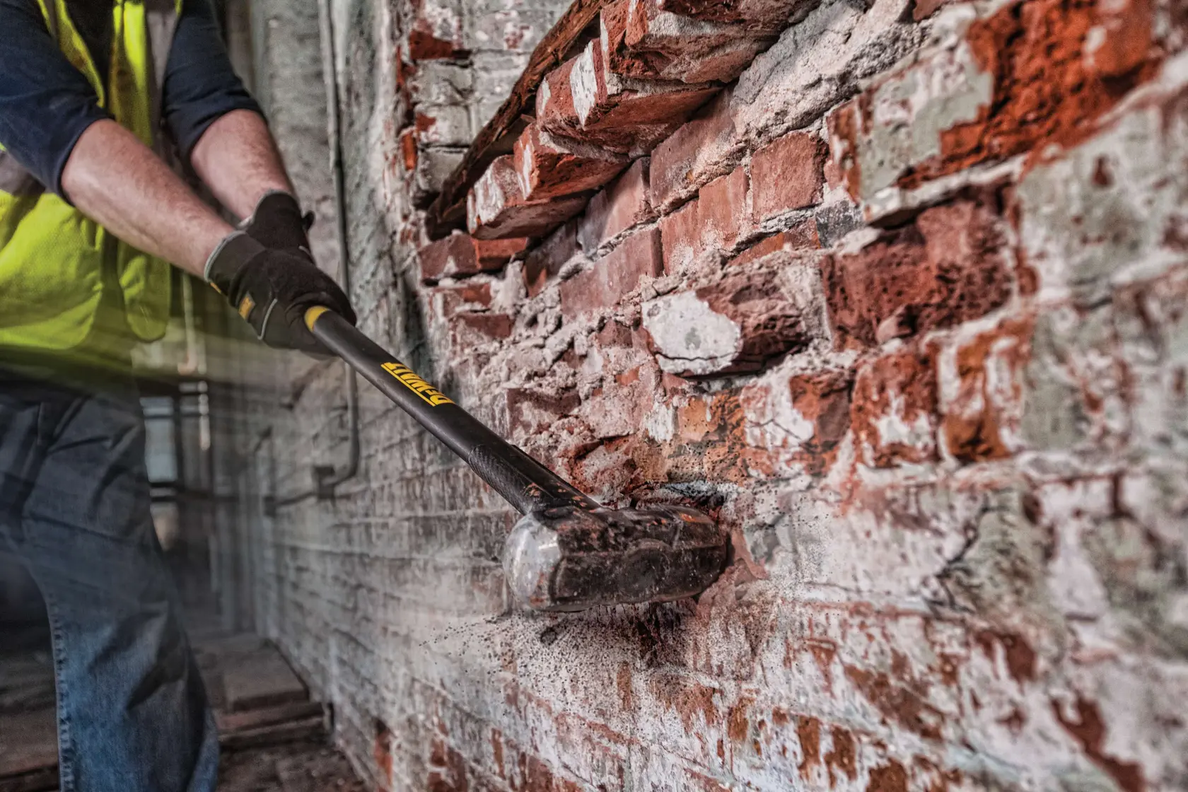 A person wearing gloves and a safety vest uses a DEWALT sledgehammer (model DWHT56029) to break a brick wall. Dust and debris scatter as the hammer strikes the bricks.