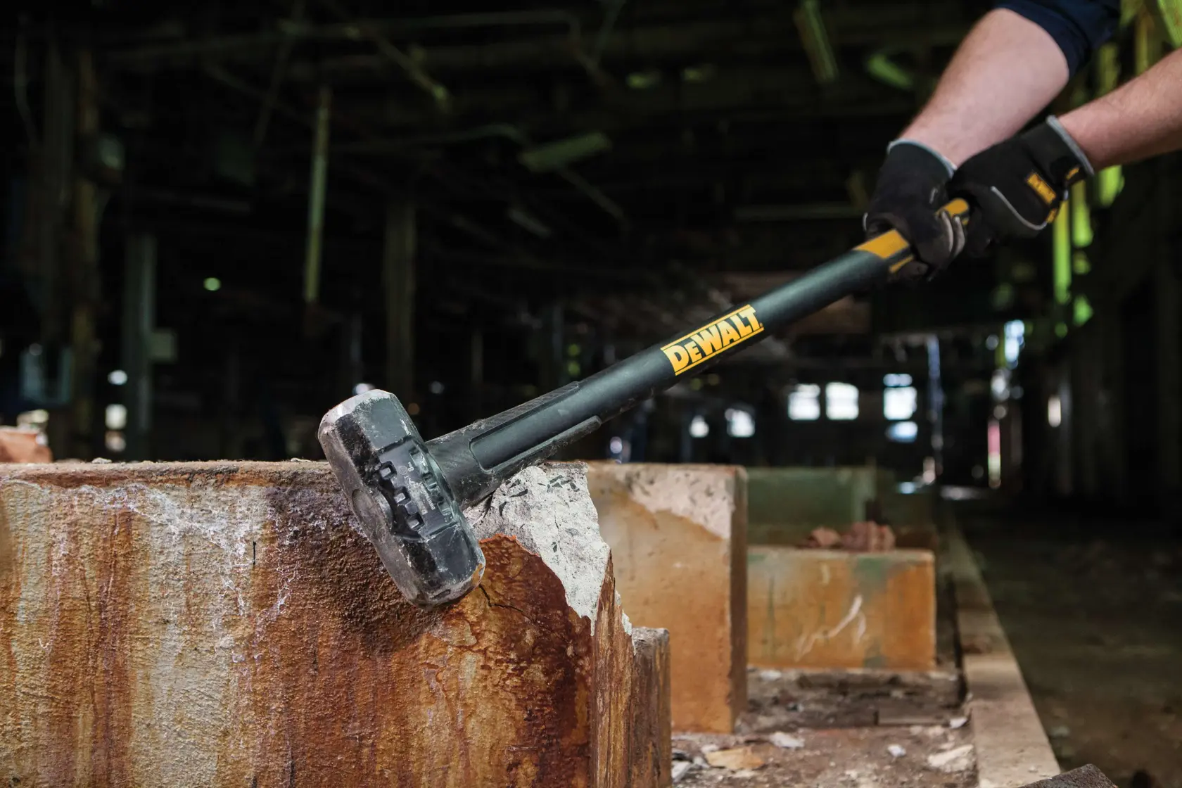 A person is using a DEWALT sledgehammer to break a concrete block in an industrial setting.