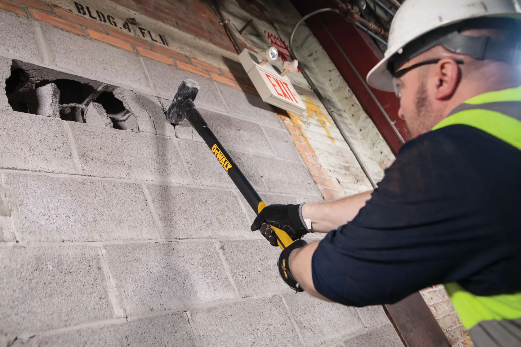 A person wearing safety gear uses a DEWALT sledgehammer (model DWHT56029) to break through a concrete block wall indoors.