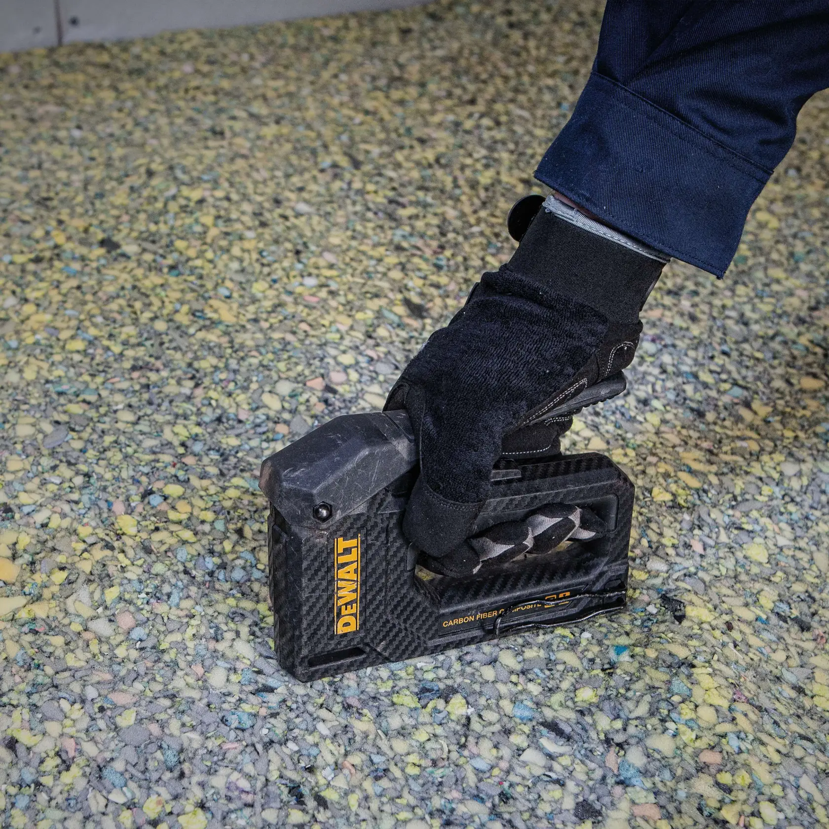 Nahaufnahme einer behandschuhten Hand, die ein DEWALT-Handwerkzeug aus Karbonfaserverbundstoff auf einer texturierten Oberfläche mit kleinen, bunten Steinen hält.