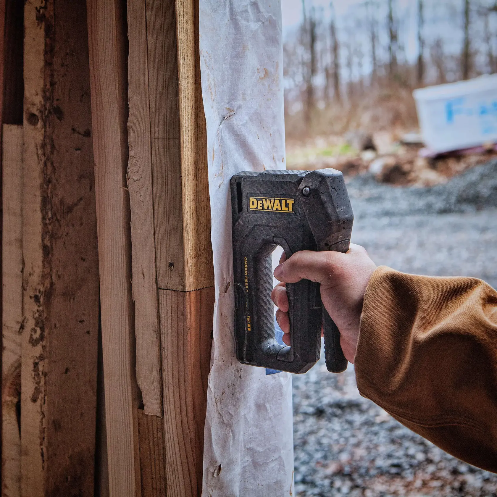Eine Person mit einem schwarzen Arbeitshandschuh verwendet einen DEWALT-Handtacker, um Isoliermaterial an einer Holzwand zu befestigen.